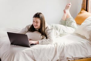 Een jonge vrouw zit op bed op haar laptop.